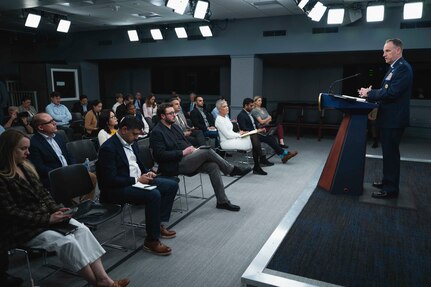A man in a military uniform stands behind a lectern and speaks to seated members of the news media.