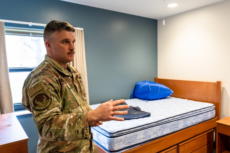 A man in a military uniform stands in front of a bare mattress and a blue wall with a window.