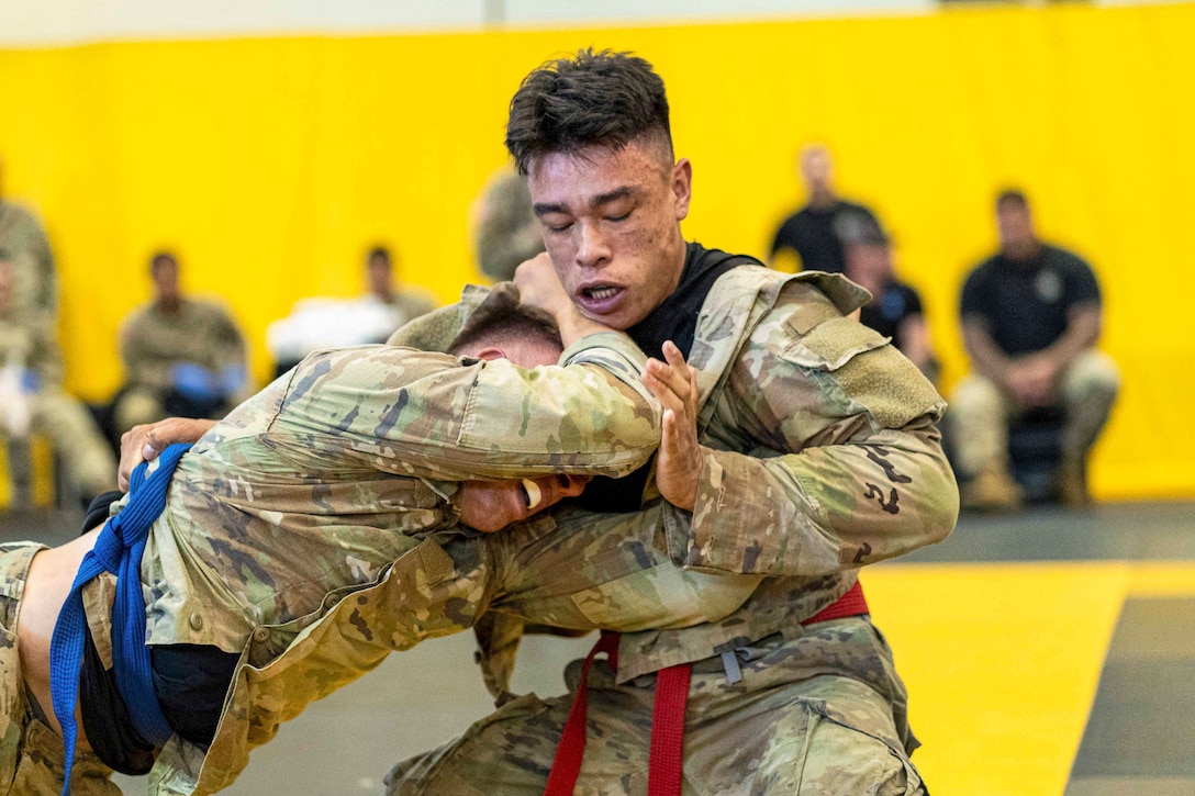 A soldier wearing a red belt wrestles a soldier wearing a blue belt in a competition as blurred soldiers watch in the background.