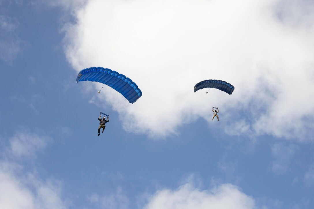 Marines free-fall in tandem with parachutes with a blue sky and clouds in the background.