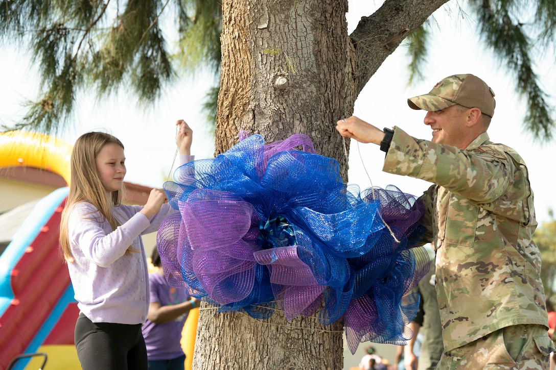 An airman and a military child tie a blue and purple ribbon on a tree with a moon bounce in the background.