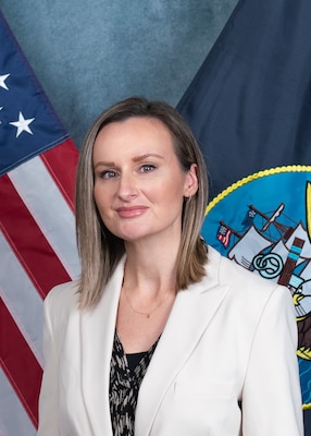 The official photo in front of the U.S. flag and the U.S. Navy flag for Dr. Vanessa Gentzen. Dr. Gentzen is wearing a white blazer and black shirt in the photo, she is sitting in front of the american and Navy flags draped behind her.