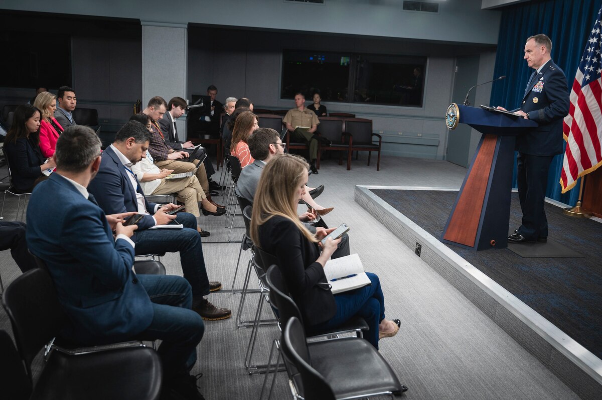 An airman in a military dress uniform stands behind a lectern addressing reporters who are seated facing the stage.