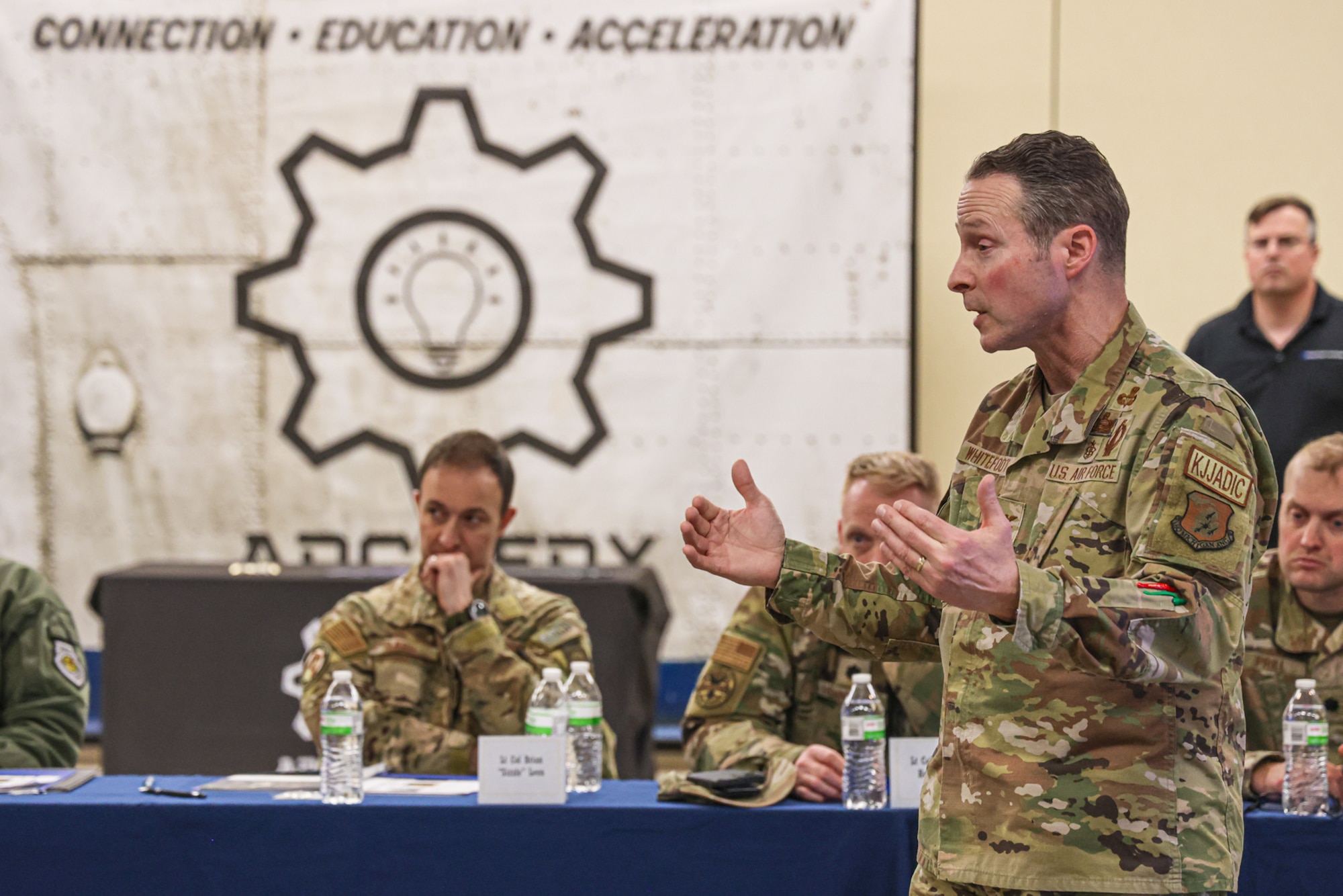 Male Air Force officer speaks while standing in room of seated military and civilian people.