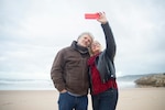 Senior couple holds up phone and smiles into camera while standing on a beach.