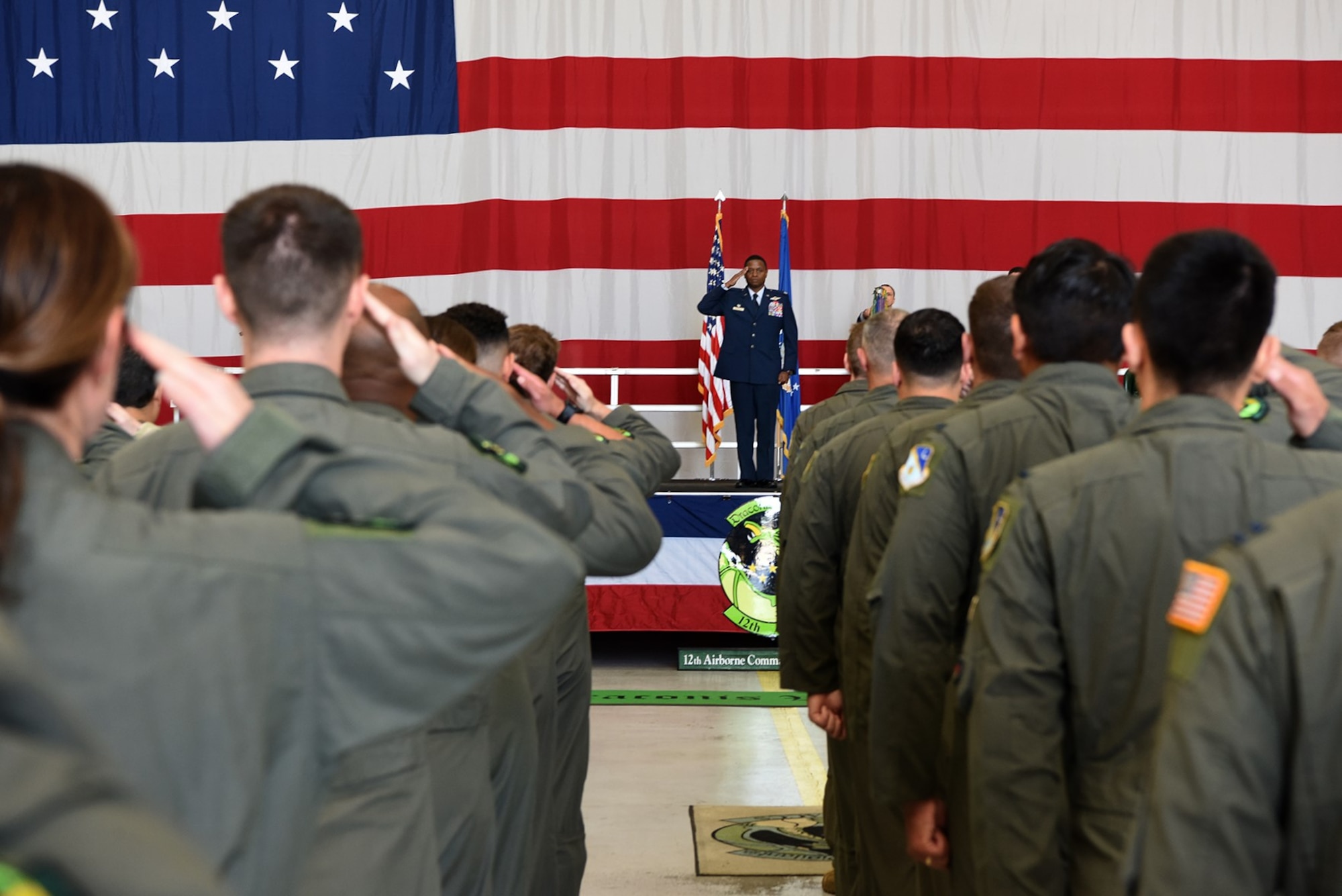 Man in military uniform standing on stage while saluting. He is placed in the middle of the photo while the backs of other military members are shown while saluting him.