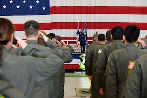 Man in military uniform standing on stage while saluting. He is placed in the middle of the photo while the backs of other military members are shown while saluting him.