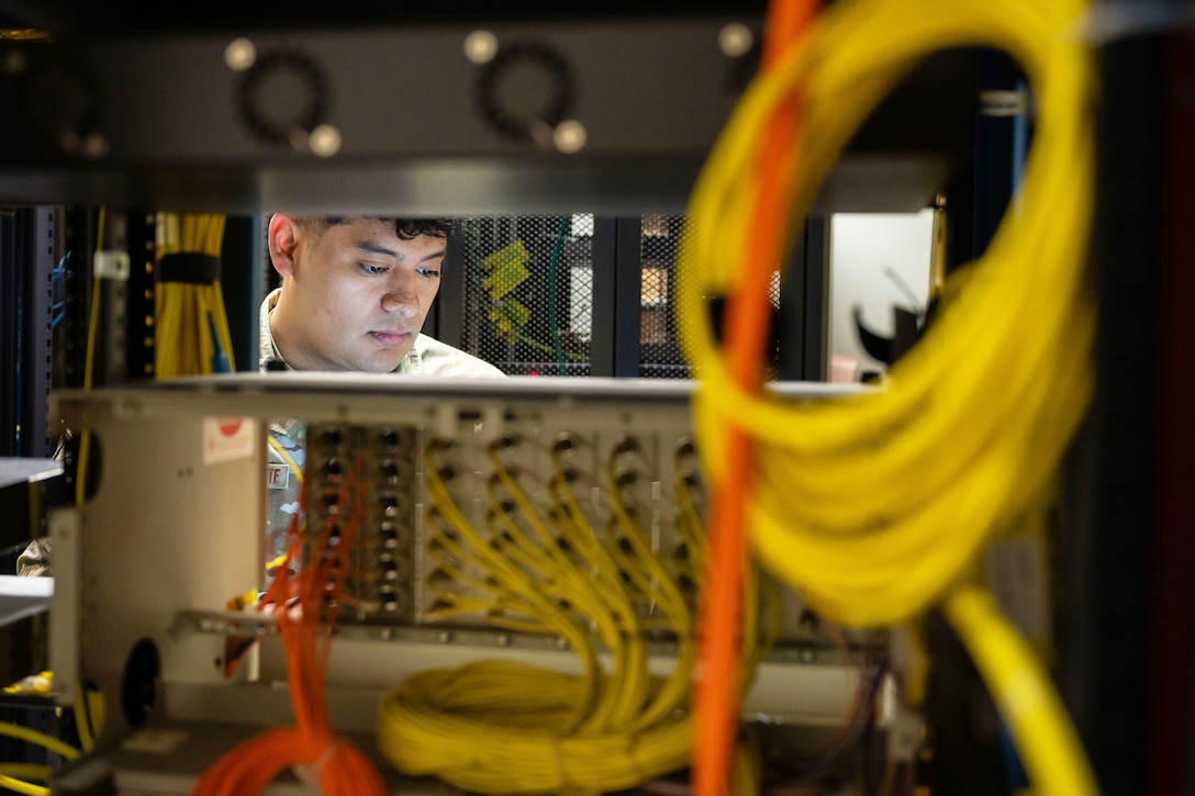 The focused face of an airman is seen through a gap in computer equipment and wiring.