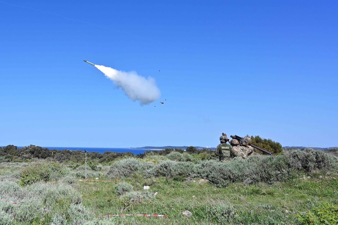 Soldiers fire a missile into a blue sky in an open area near a coastline.