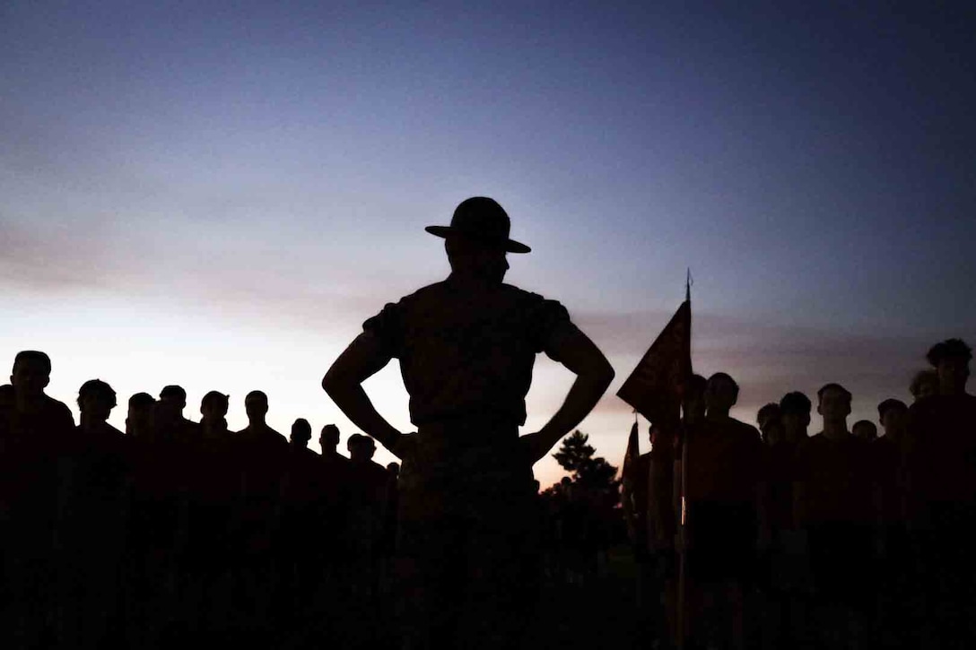 A drill instructor and a large group of people are silhouetted against the sky.