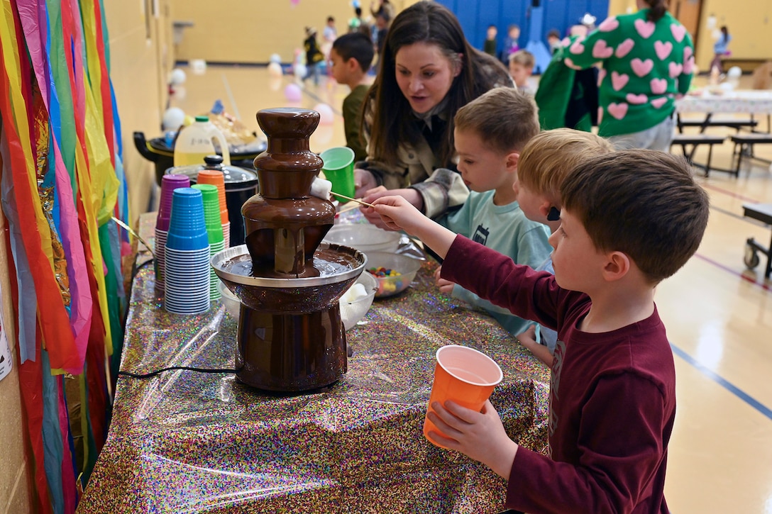 A child dips a marshmallow into a chocolate fountain as two others watch an adult doing the same.