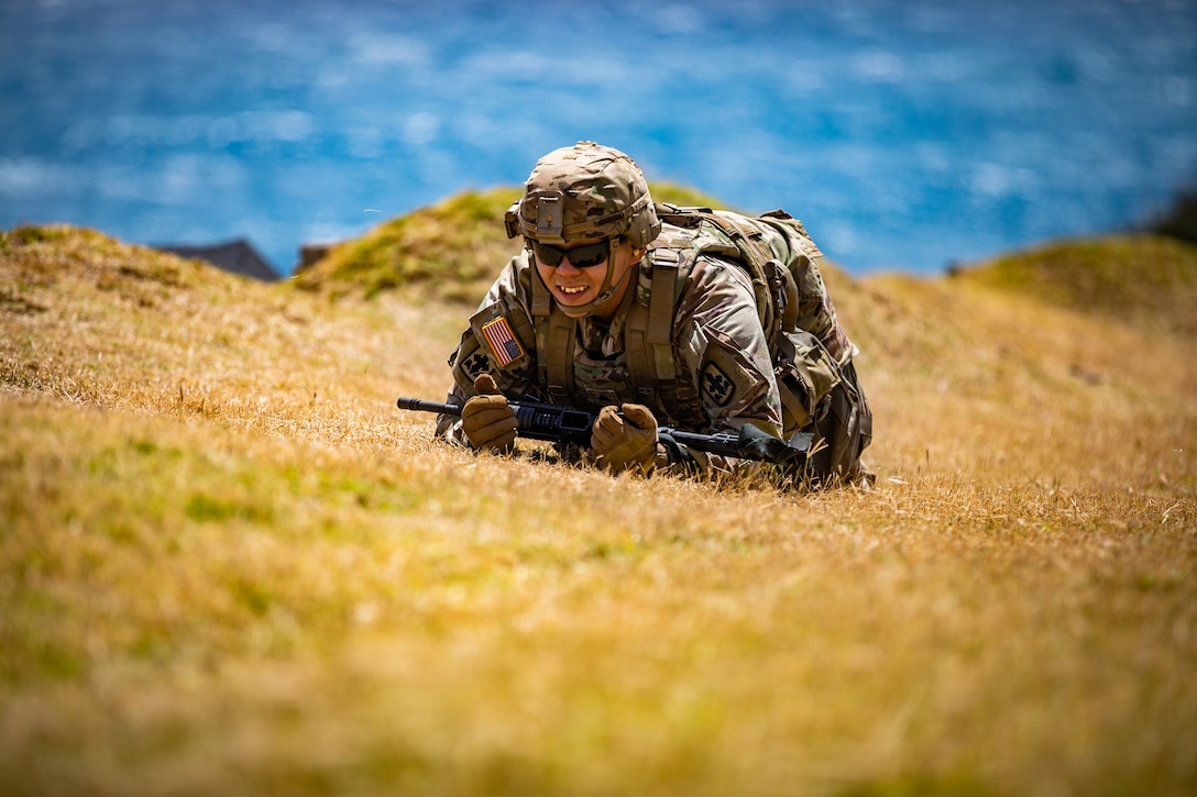 A uniformed soldier wearing a helmet and sunglasses and carrying a weapon crawls up a hill during daylight.