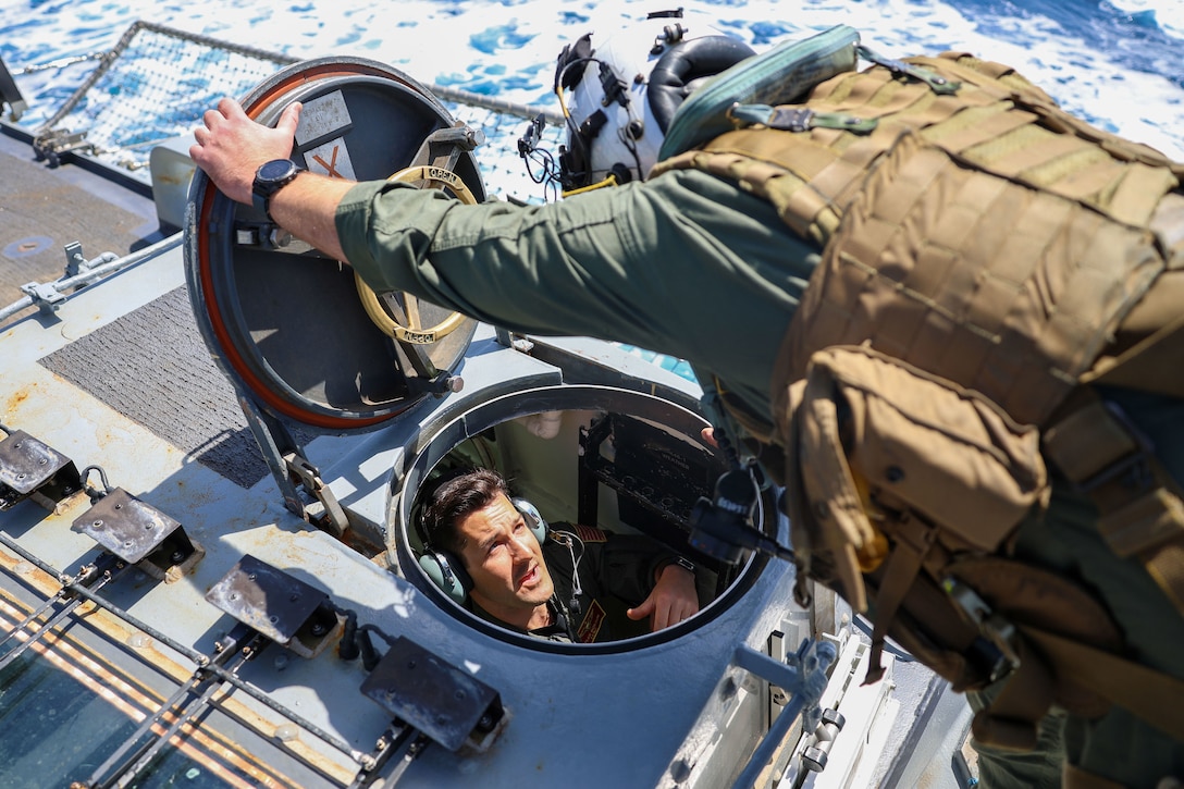 A sailor leans forward and holds open a portal to speak to another sailor below the flight deck of a ship at sea.