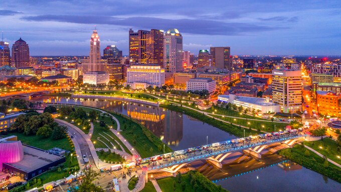 A photo of a city skyline as seen from above showing buildings, bridges, a river all aglow in a myriad of colored lights. There is a large swath of greenspace on either side of the river.