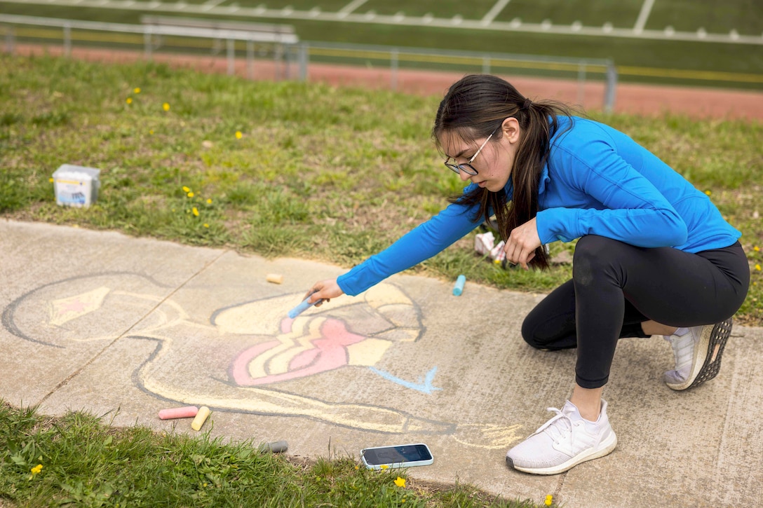 A Marine uses chalk to draw Wonder Woman on a sidewalk next to a field.