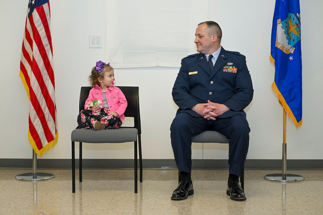 An airman and his young daughter lock eyes while sitting next to each other in between two flagpoles.
