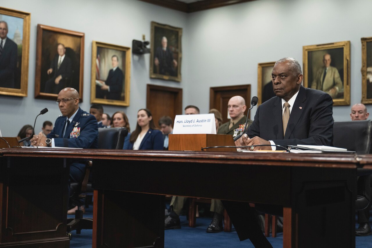Secretary of Defense Lloyd J. Austin III and Joint Chiefs Chairman Air Force Gen. CQ Brown, Jr., sit at a long table in front of microphones.
