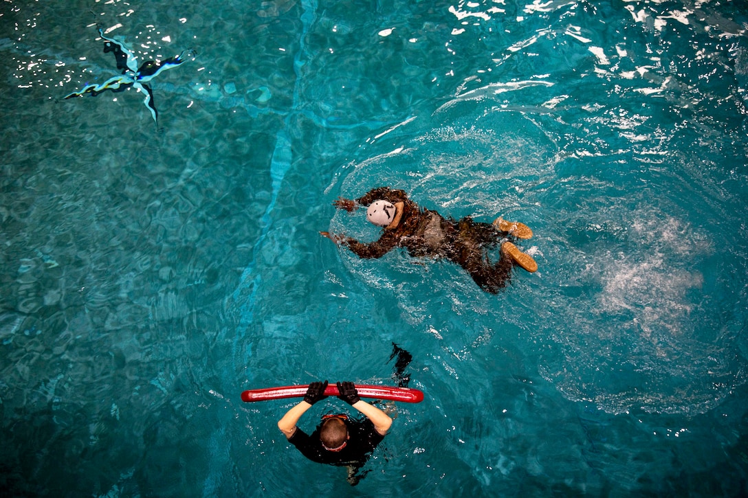 A Marine wearing a helmet, equipment and shoes swims partially underwater in a pool while another Marine stands close by holding a thin red raft.