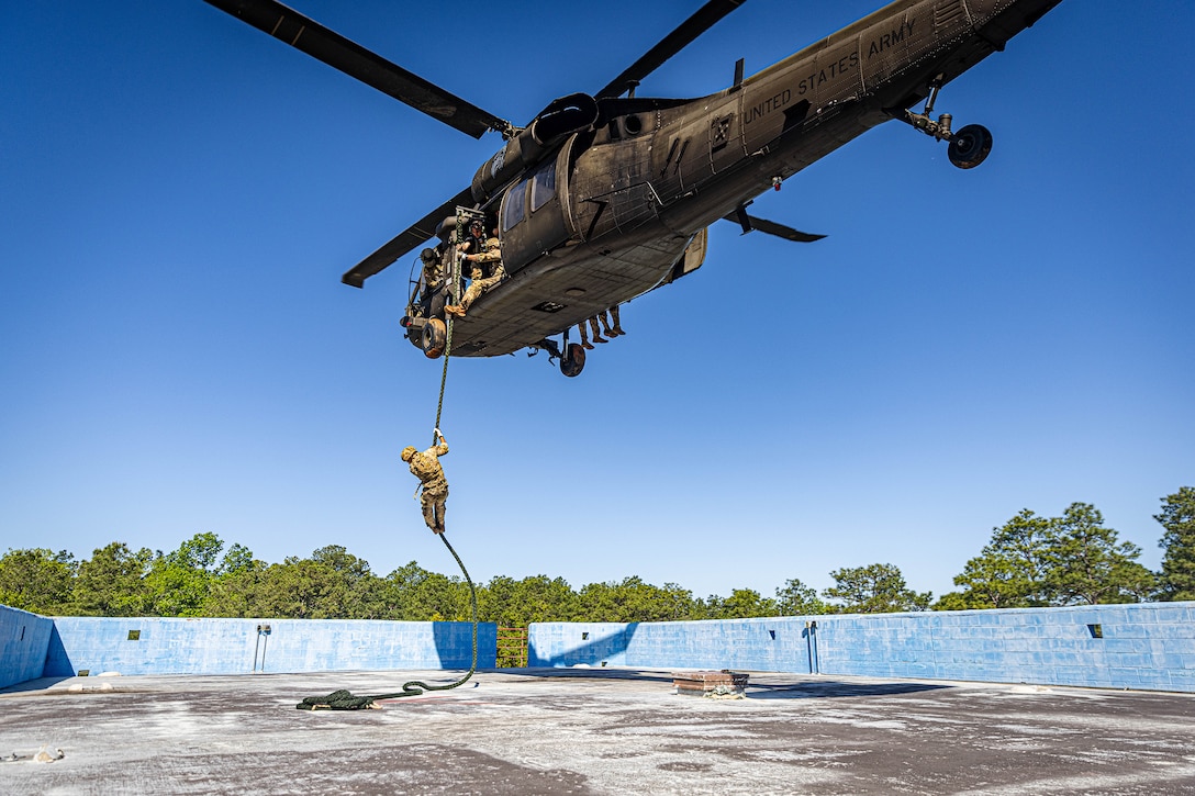 A soldier fast-ropes from a military helicopter onto a roof as others watch him from inside the aircraft during daylight.