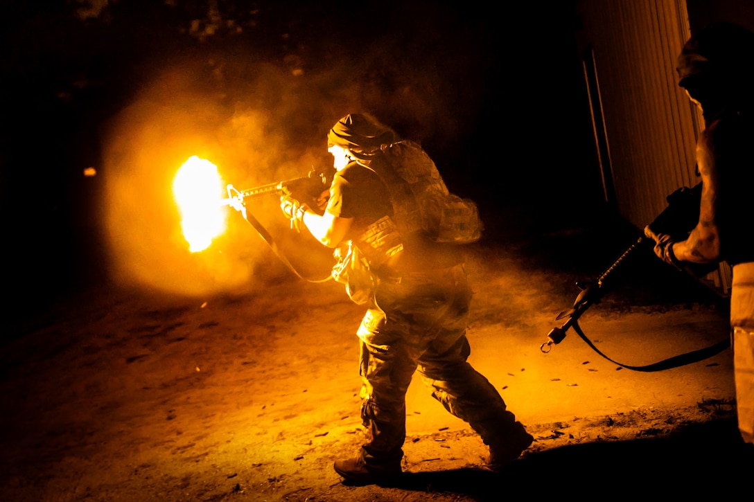 Two soldiers hold weapons as they approach opponents during night training. A bright orange flame can be seen emitting from the first soldier's weapon.