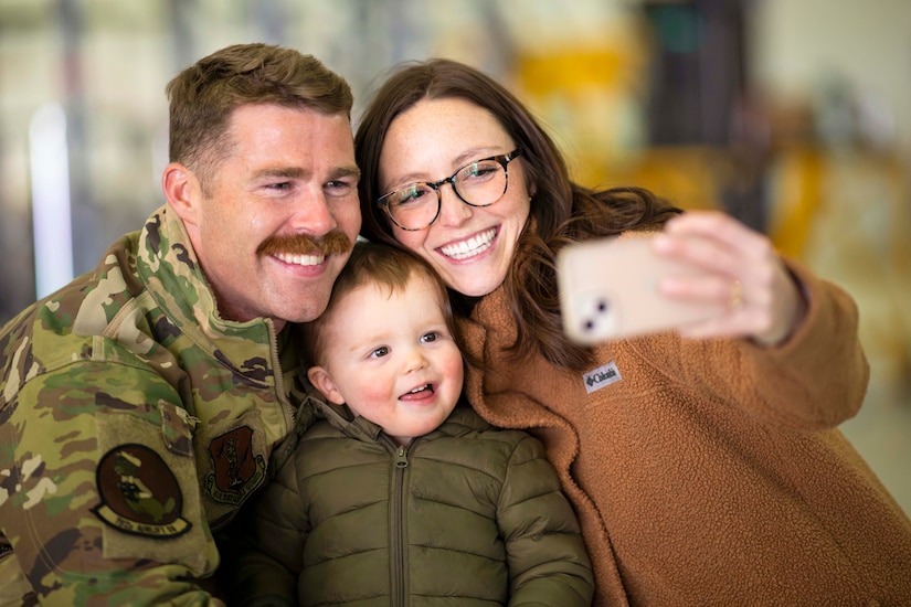 A person holds a phone horizontally to take a picture with a child and an airman.