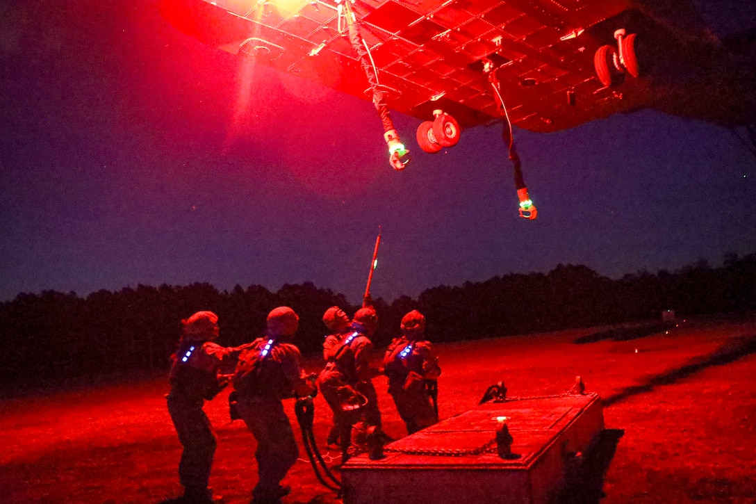 Marines attempt to connect an object to a hovering helicopter partially seen from above as it illuminates red light.