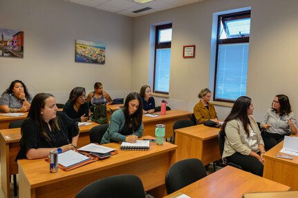 Civilians take notes while seated at desks in a classroom.