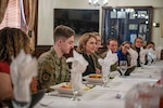 A woman in civilian attire sits at a table with service members in uniform.