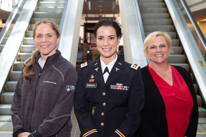 Three women, one in military uniform, pose for a photo in front of escalators.