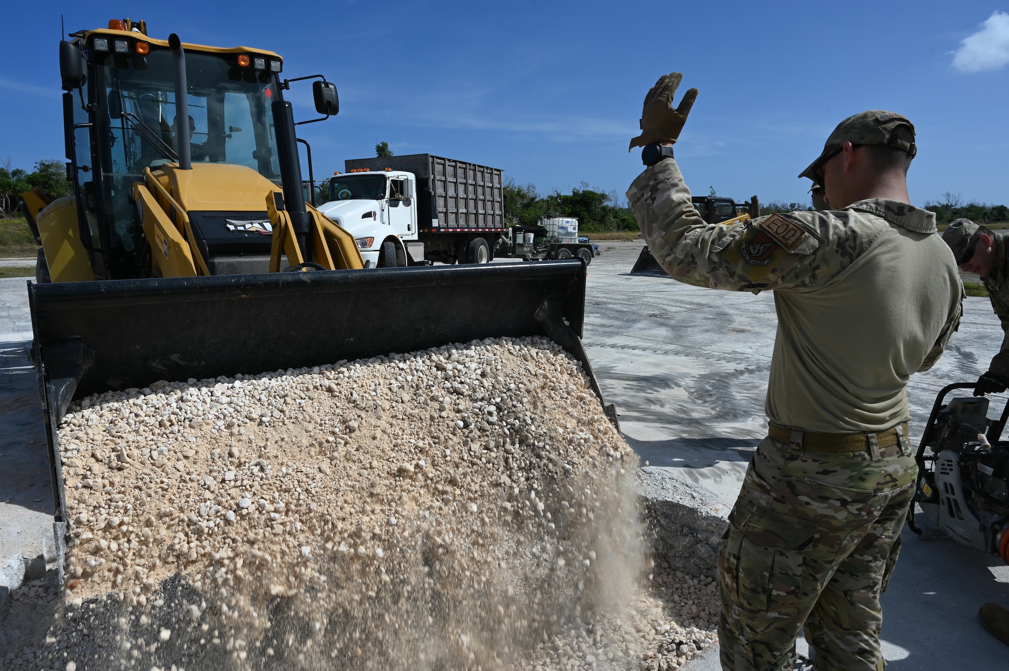 An Airman directs another Airman who is operating a backhoe on disposing crushed stones on a crater.