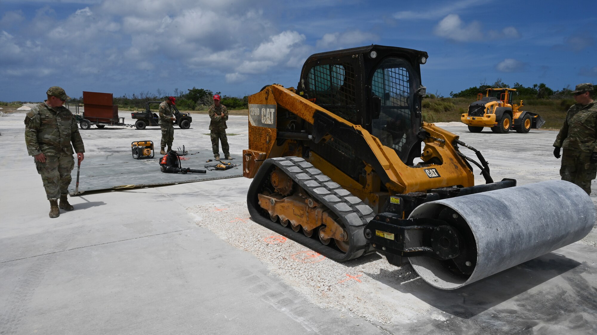 Airmen use a compact track loader to pull a fiber reinforced polymer matting over a crushed stone crater repair.