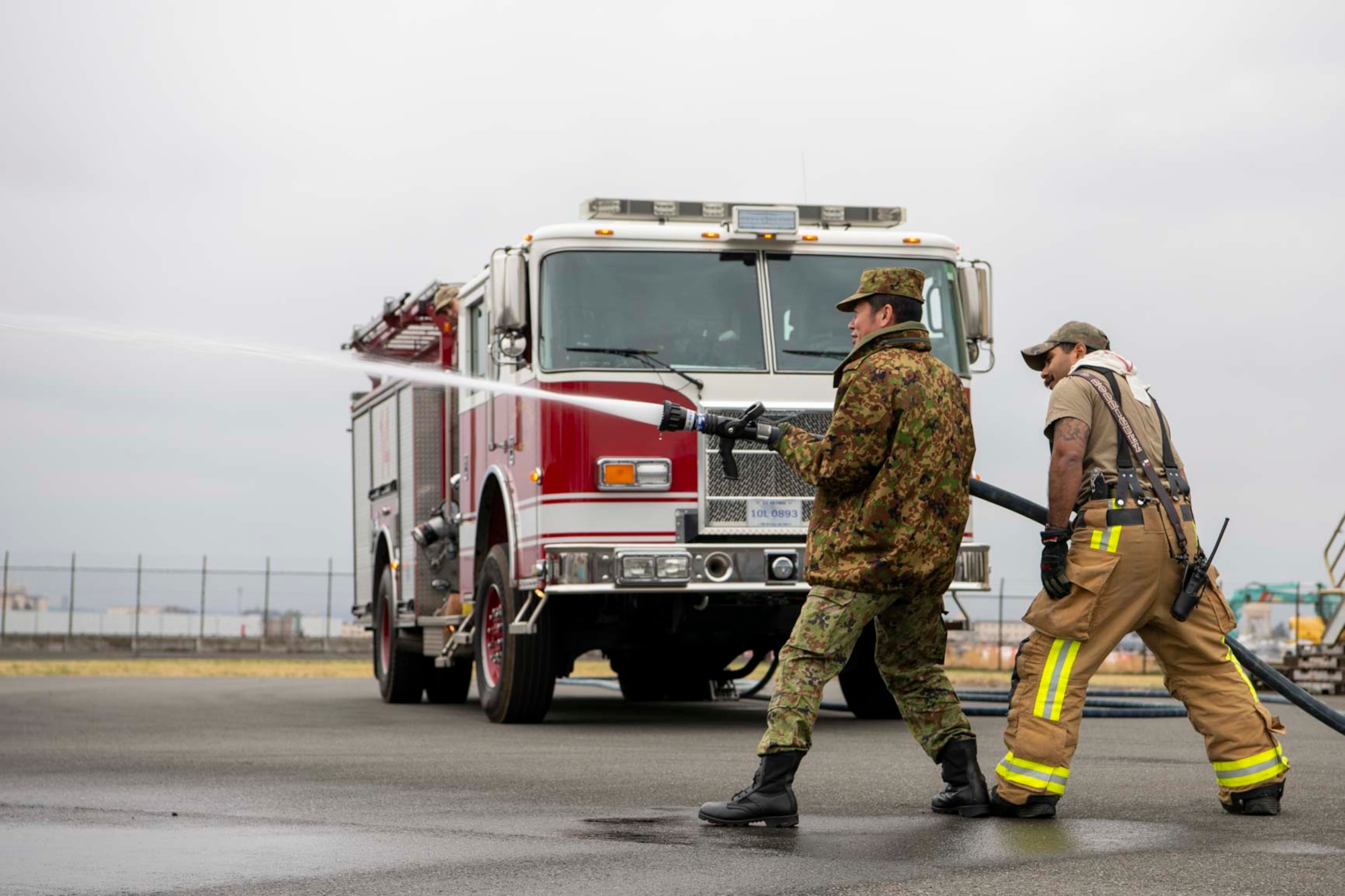 A Japanese military member and U.S. firefighter spray water out of a fire hose.