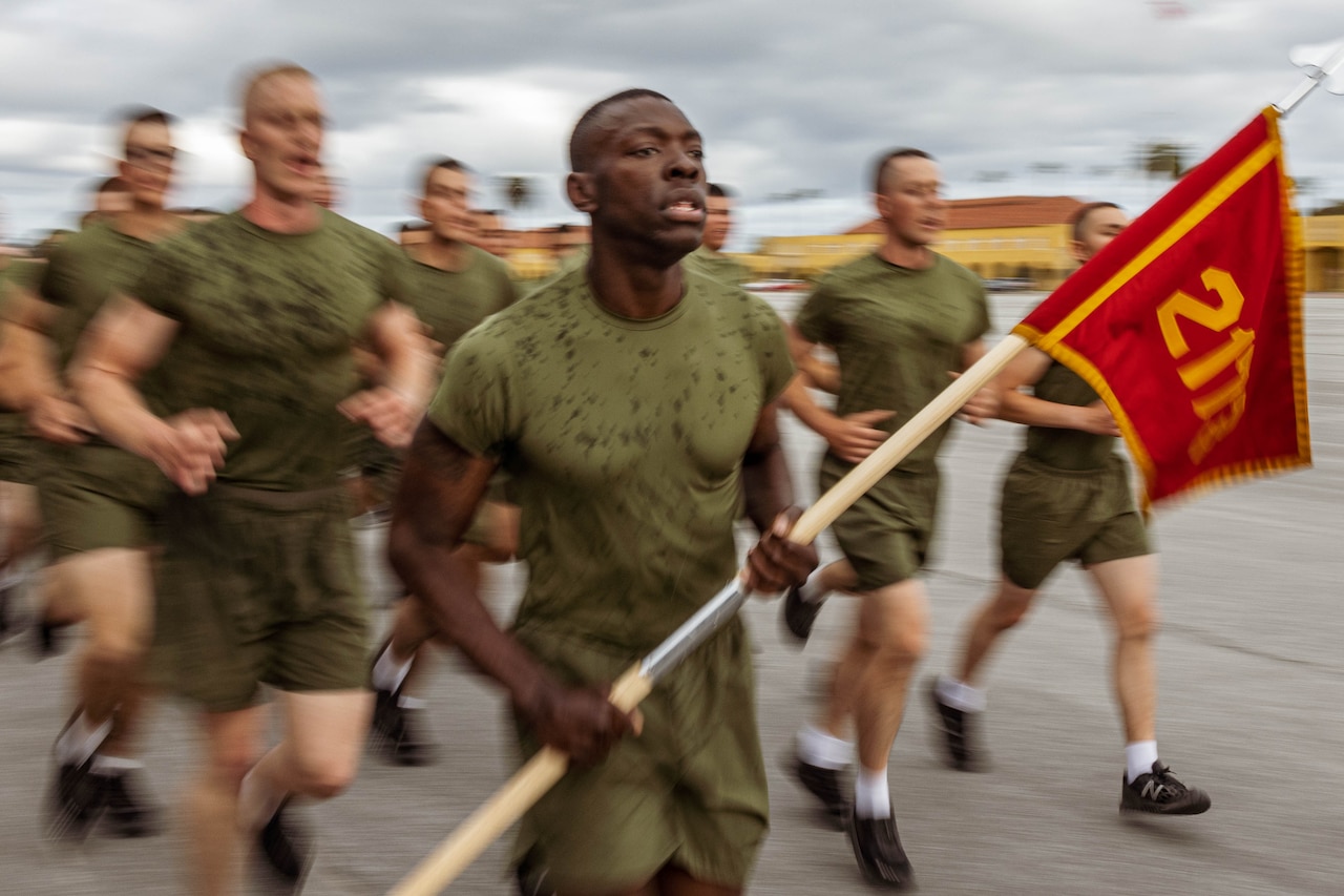 A Marine Corps recruit carrying a flag leads fellow recruits while running.