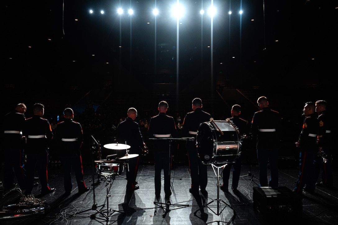 Military musicians stand on stage under bright lights while facing the audience. Drums can be seen on the stage.
