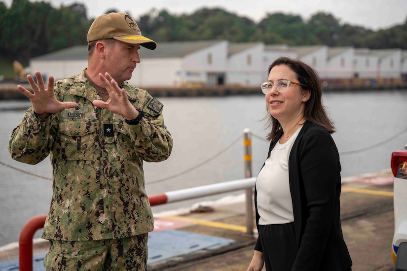 A person in civilian clothes talks with a uniformed service member. They are near a shoreline aboard a boat.