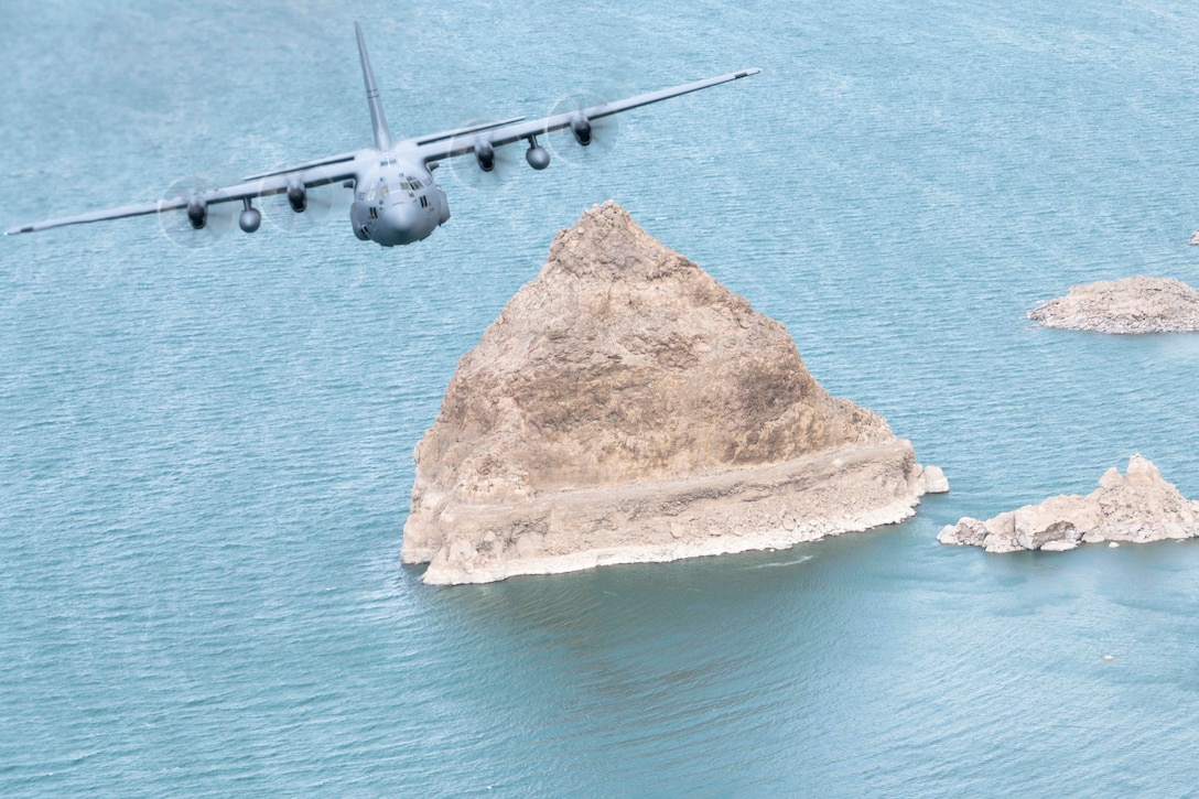 An aircraft flies above a large rock in a body of water with smaller rocks to its right.