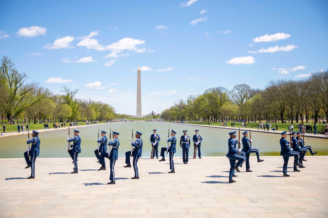 A airmen in ceremonial uniforms march while carrying rifles in front of the reflecting pool with the monument in the background.