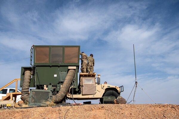 Three people stand on the roof of the cab of a Humvee to secure a panel to the vertical extension from the bed of the truck.