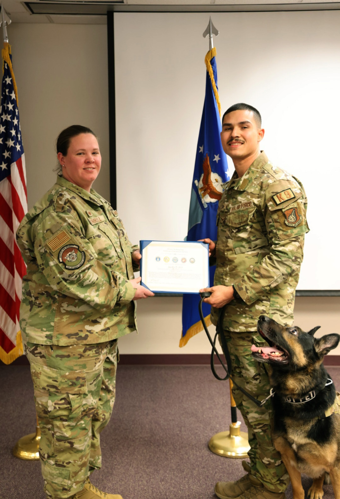 Two security forces members hold a certificate while MWD Jocky looks up and smiles.