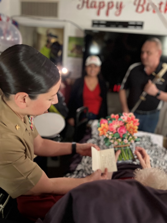 U.S. Marine Corps Major Shannon Potts, A Manpower Officer assigned to Headquarters Company, 23rd Marine Regiment, 4th Marine Division presents a birthday card to retired U.S. Marine Corps Corporal Lou 'Mama Lou' Keller for her years of service during Mama Lou's 99th Birthday Celebration, March 30, 2024, in San Jose, CA. Mama Lou was stationed at the Pentagon during the height of WWII processing Purple Hearts.