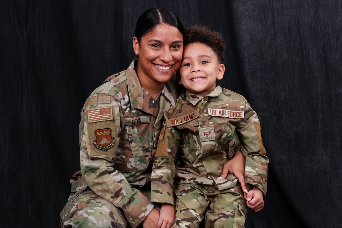 An airman smiles for a photo with her son against a black background; both wear camouflage uniforms.