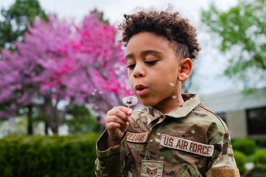 A child wearing a camouflage outfit blows on a dandelion outside, with pinkish-purple flowering spring trees in the background.
