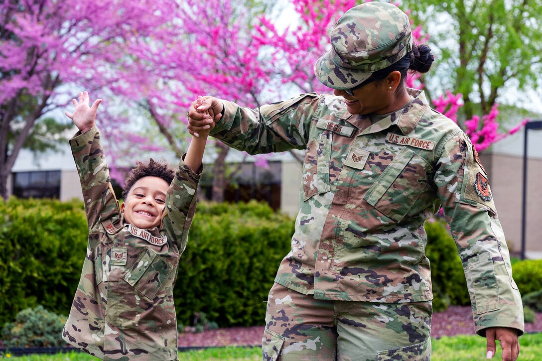 An airman holds hands with son outdoors as he raises his arms and she lifts him; both wear camouflage uniforms.