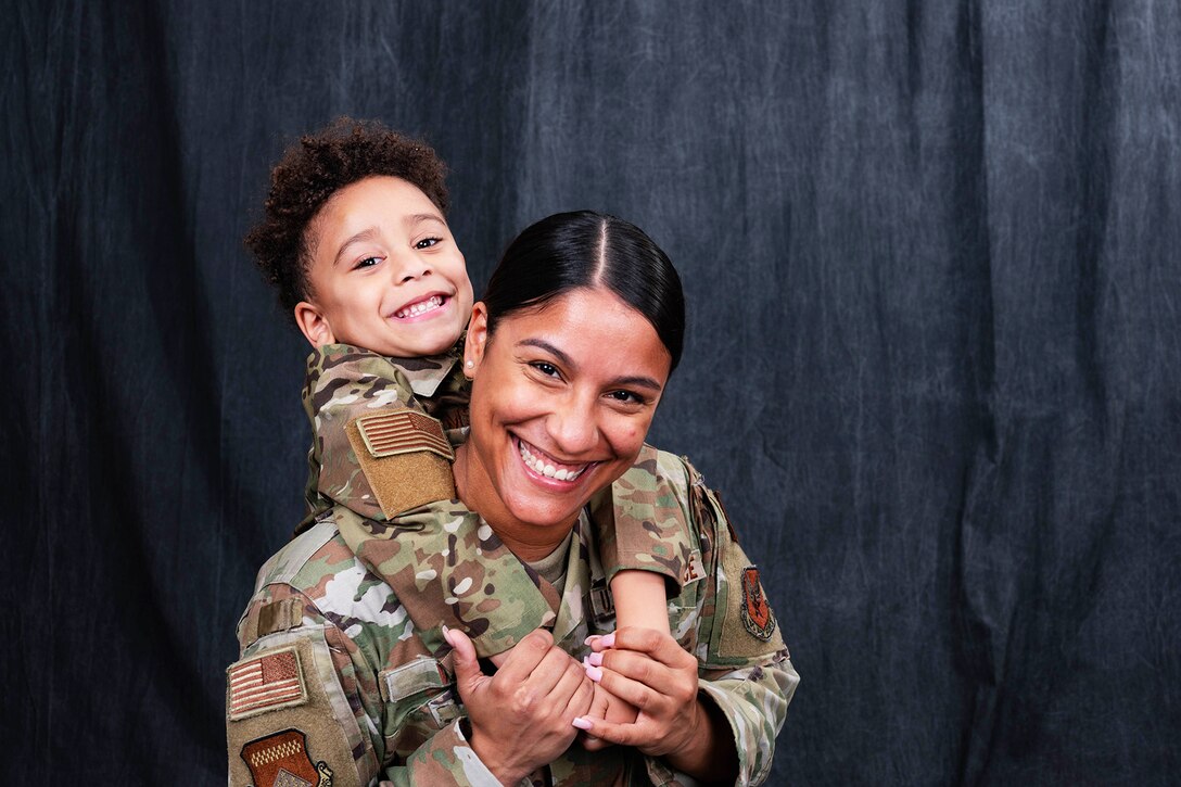 A child wraps his arms around his smiling mom's shoulders while smiling behind her; both wear camouflage uniforms.