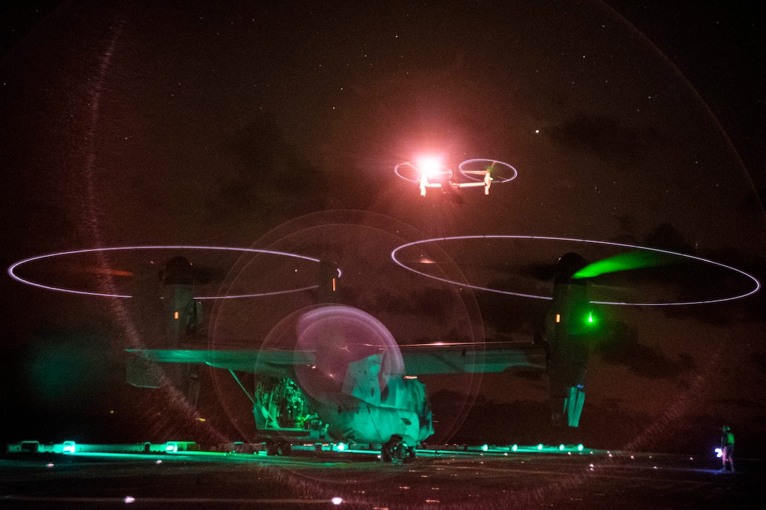 Lights of two Osprey aircraft and their propellers are seen on a ship’s deck and in the dark sky.