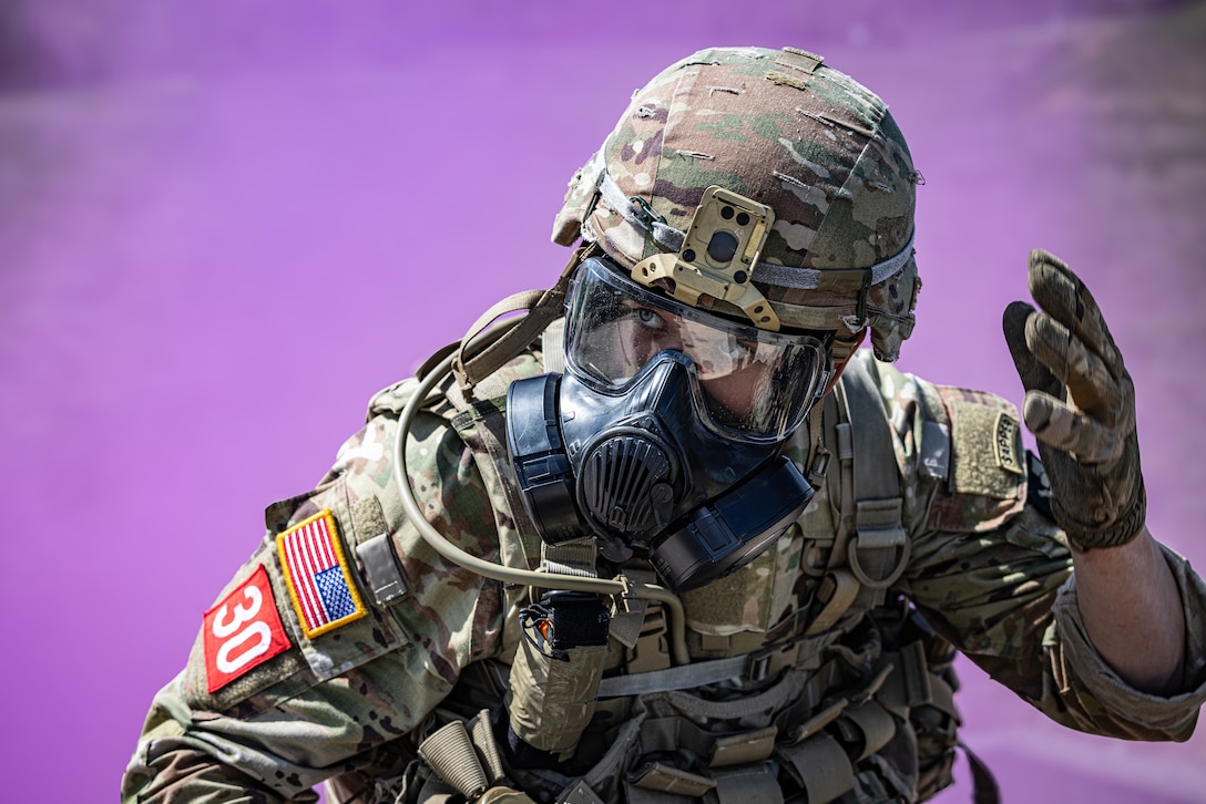 A uniformed soldier in a helmet, goggles and mask gestures as purple smoke is visible in the background.