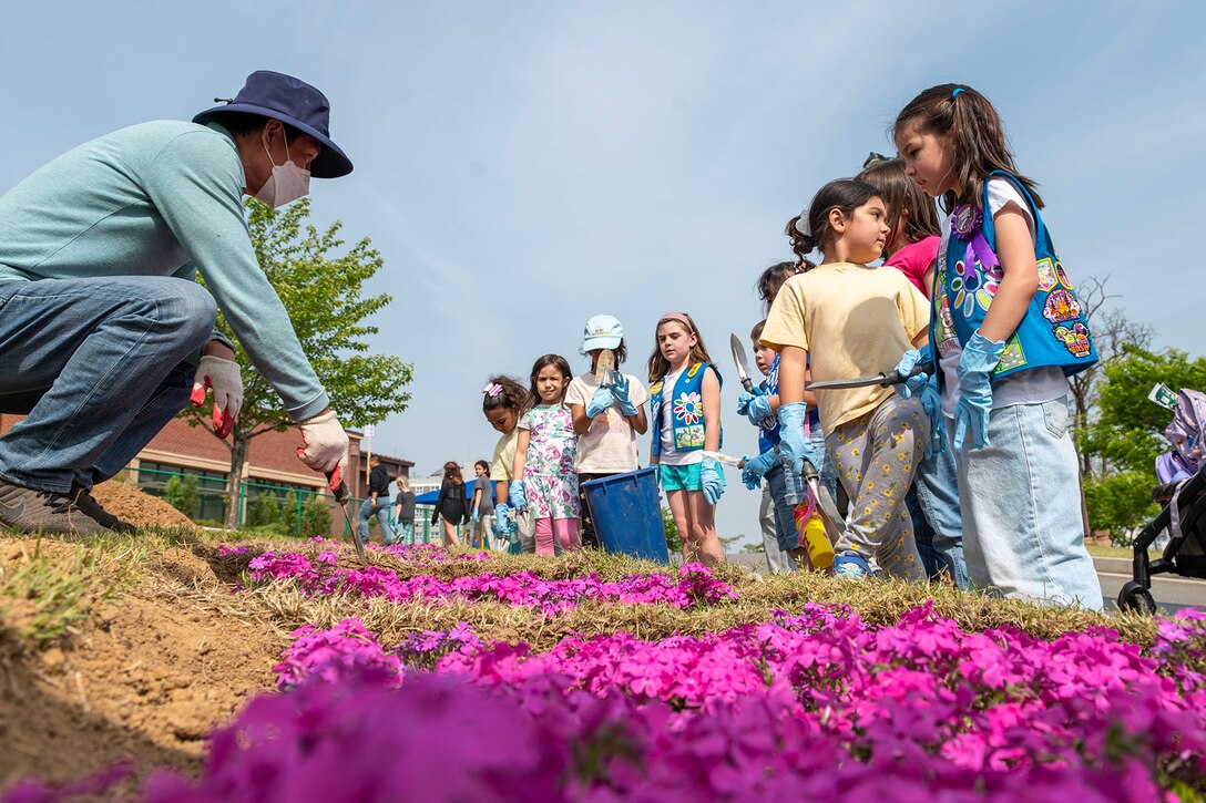 A group of children hold gardening tools in preparation for planting vegetation as an adult squats and gestures toward the ground with a tool. Purple flowers are visible in the foreground.