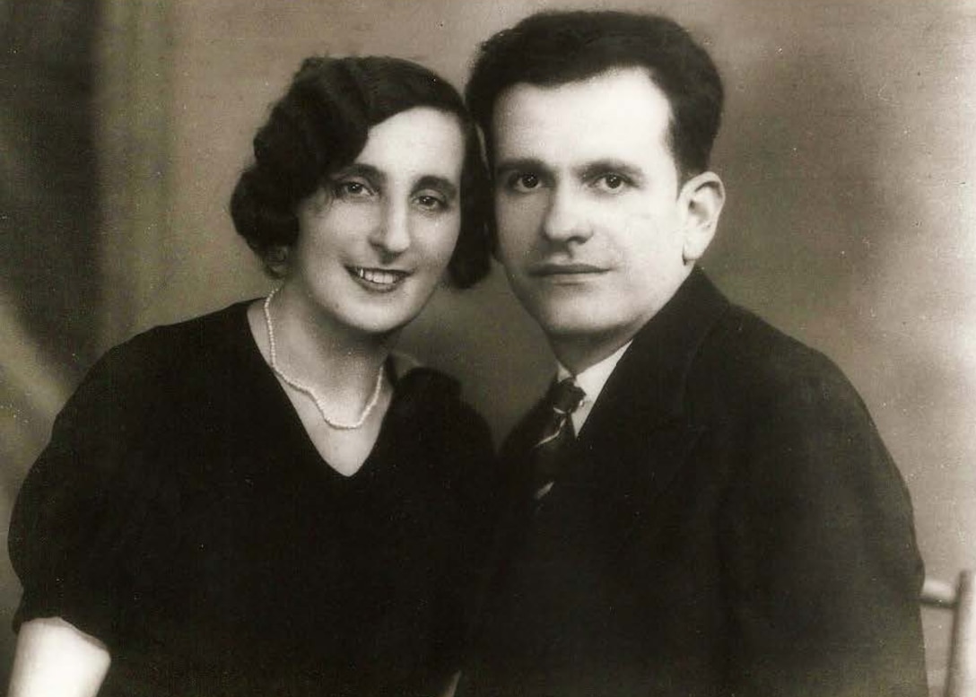 Historical photo of two people in dark clothing smiling at the camera. The woman has dark hair and is wearing a pearl necklace and the man is in a dark suit and also has dark hair.