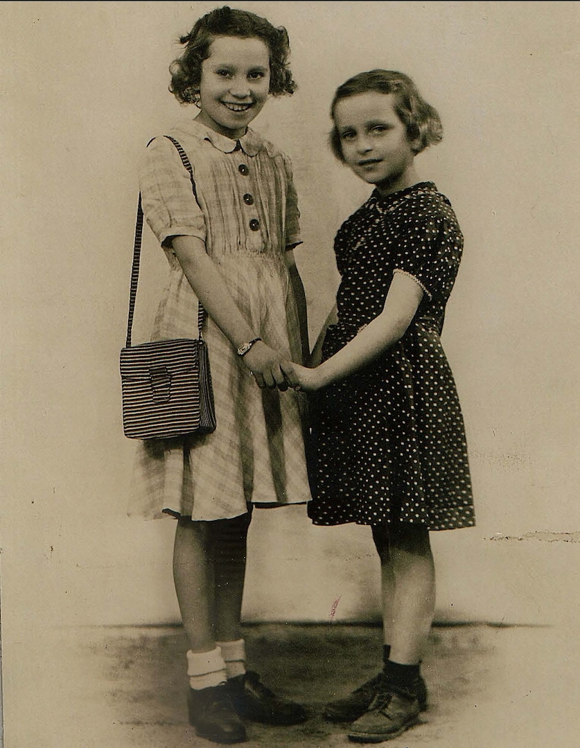A historical  black and white photo of two children holding hands in patterned dresses. Both are smiling. One is wearing a purse over her shoulder. They are about six and seven years old.