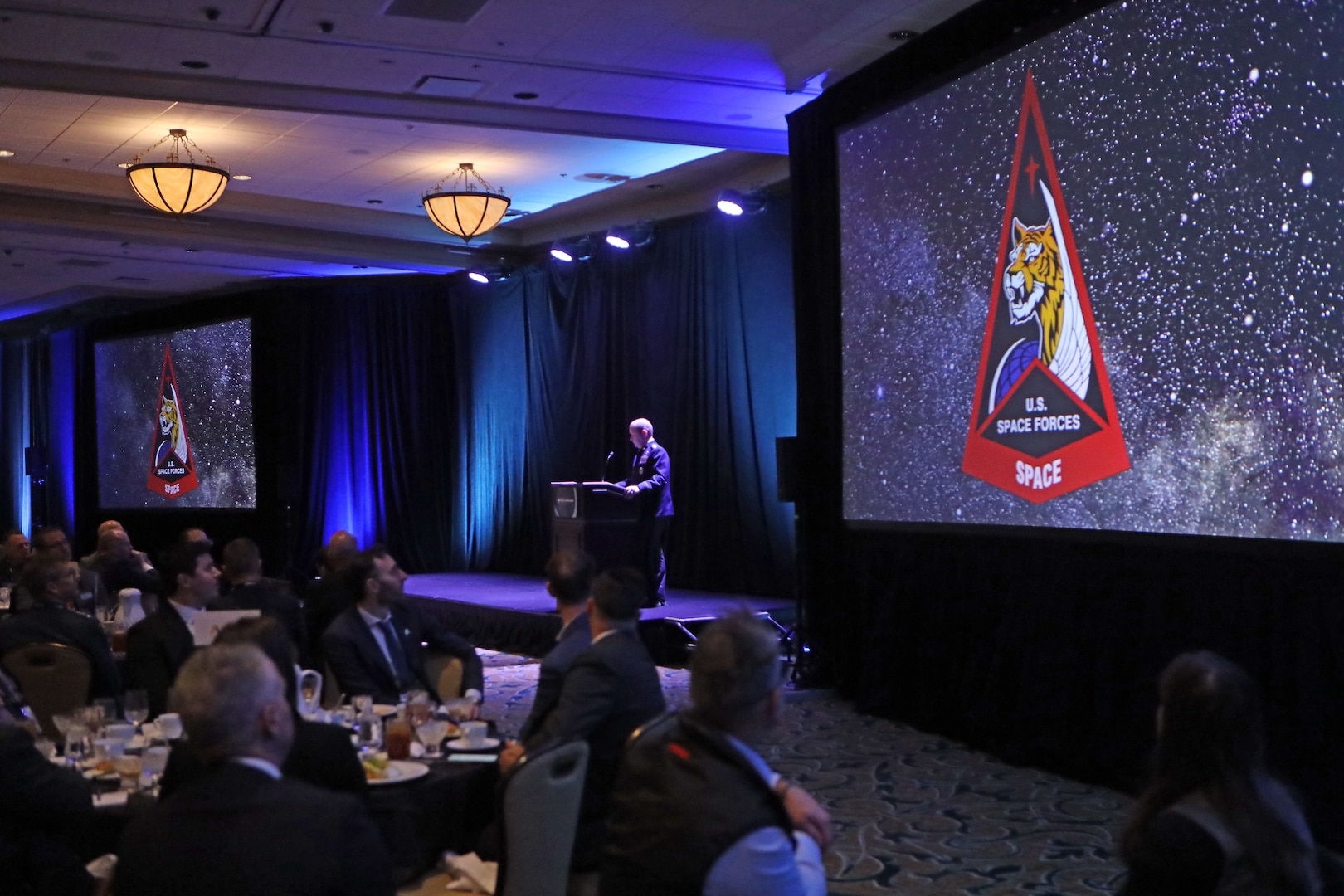 man speaking at podium next to screen projector